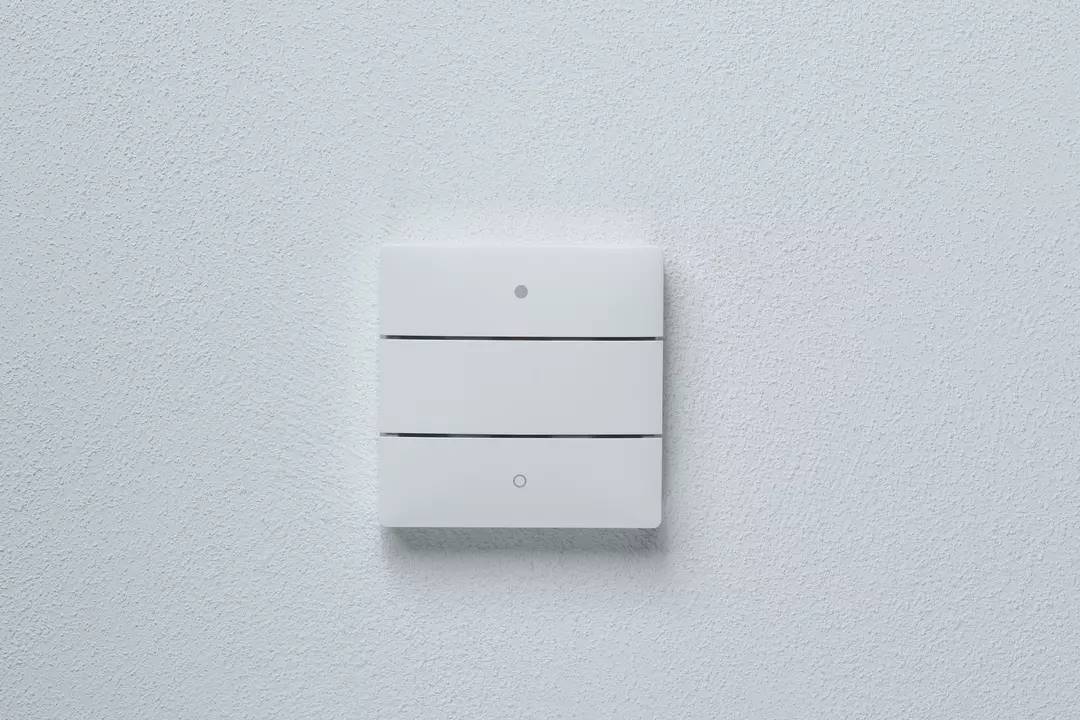 Ein weißer Lichtschalter mit drei horizontalen Tasten an einer weißen Wand.