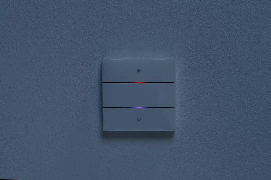 Ein grauer Wandschalter mit drei horizontalen Tasten und einer roten Kontrollleuchte; minimalistisches Design auf grauer Wand.