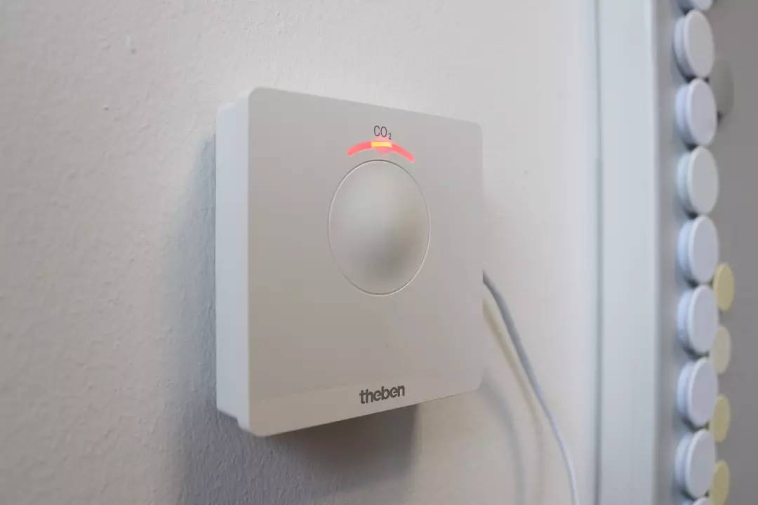 Ein weißer CO2-Sensor mit roter Lichtanzeige, montiert an einer Wand, zeigt erhöhte CO2-Werte an. Ein Kabel führt zum Gerät.