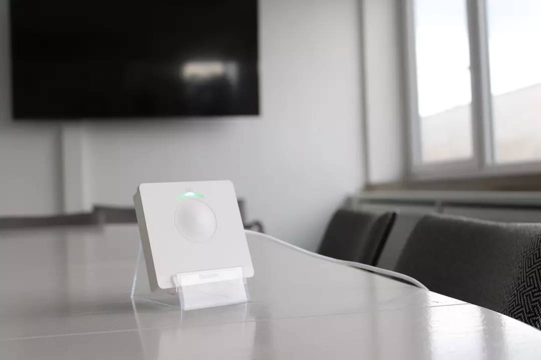 Ein rechteckiges Gerät mit einem grünen Leuchtsignal steht auf einem Tisch in einem Konferenzraum mit einem Bildschirm im Hintergrund.