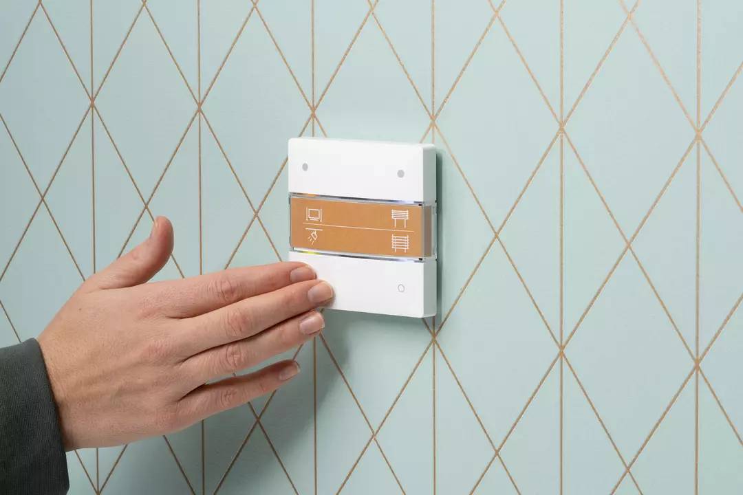 Eine Hand berührt ein modernes Wandpaneel mit Symbolen für Lichtsteuerung, auf einer geometrisch gemusterten, hellblauen Wand.