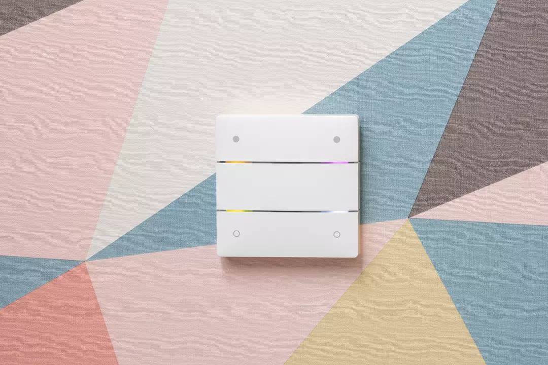 Ein moderner dreifacher Lichtschalter an einer farbenfrohen, geometrischen Wand mit Dreiecksmuster in Rosa, Blau, und Grau.