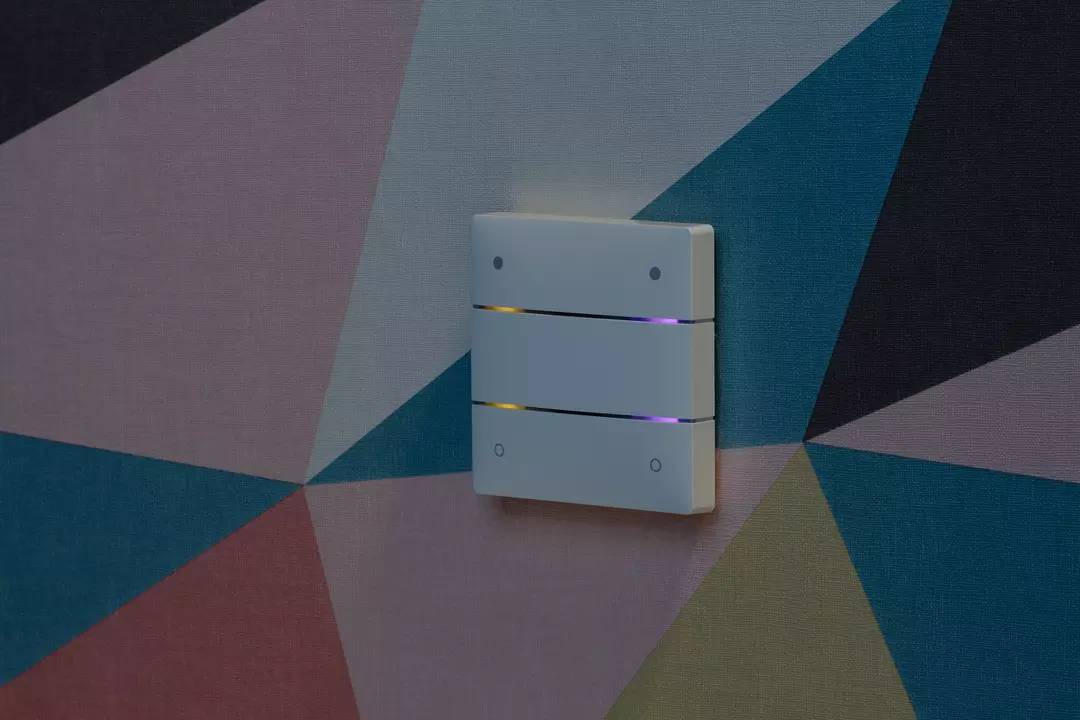 Ein dreiteiliger Wandschalter auf bunter, geometrischer Tapete. Die Schalter leuchten dezent und sind vertikal angeordnet.
