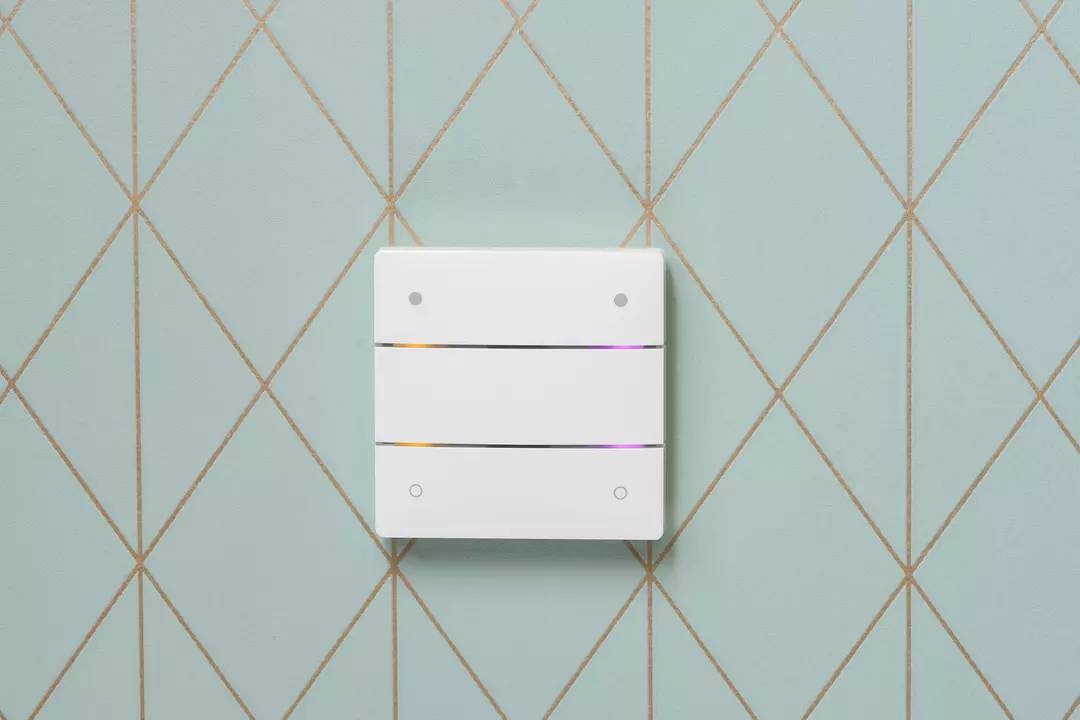 Ein weißer Lichtschalter mit drei Tasten an einer Wand mit einem geometrischen Gittermuster. Die Tasten leuchten dezent.