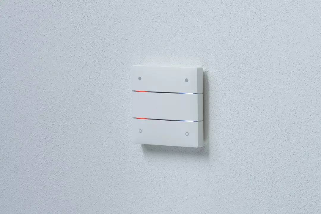 Ein weißes Wandgerät mit horizontalen Schlitzen, aus denen schwaches, blaues und rotes Licht leuchtet, montiert an einer weißen Wand.