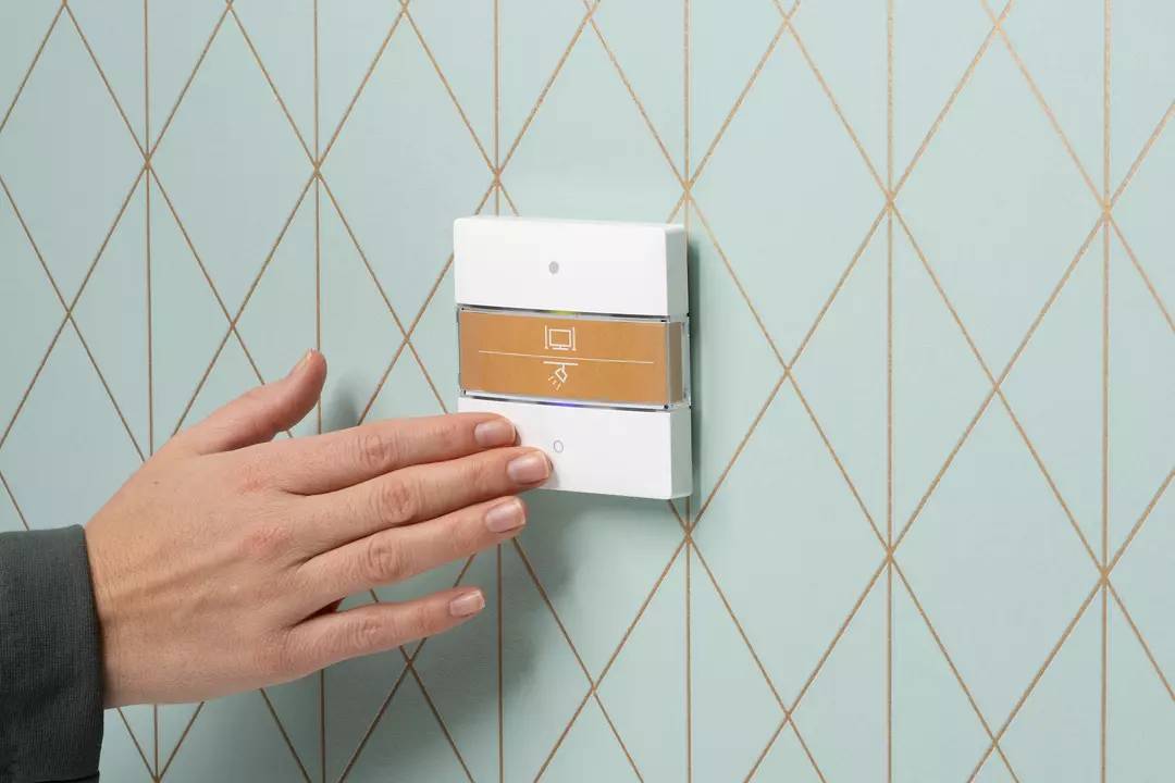 Hand berührt einen modernen Lichtschalter an einer hellblauen Wand mit goldenen Linien. Der Schalter hat eine grafische Anzeige.