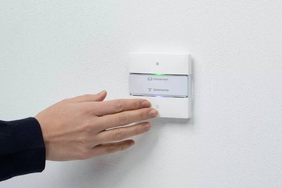 Eine Hand berührt einen Lichtschalter an der Wand. Der Schalter hat zwei Knöpfe, beschriftet mit 'Entfernen' und 'Deckenleuchte'.