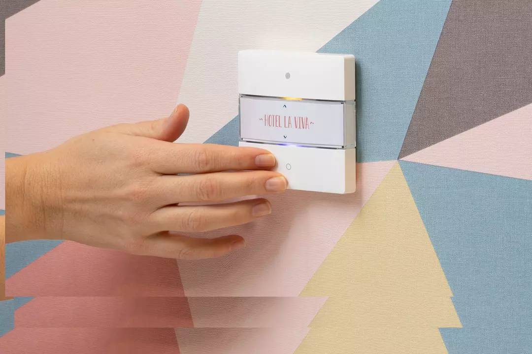 Eine Hand nähert sich einem modernen Lichtschalter an einer Wand mit geometrischem Muster und der Aufschrift 'Hotel La Viva'.