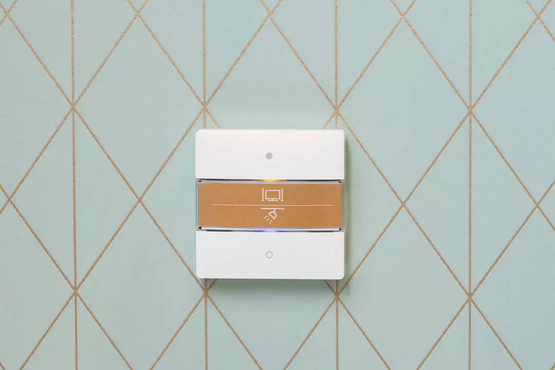 Ein weißer Lichtschalter auf einer hellblauen Rautenmuster-Wand. Der Schalter zeigt ein Symbol für ein Licht.