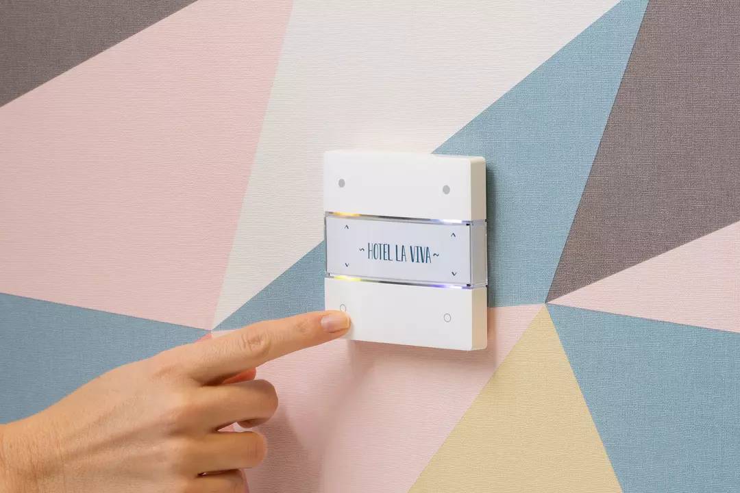 Eine Hand berührt einen modernen Lichtschalter an der Wand, auf dem 'Hotel La Viva' steht. Die Wand hat ein geometrisches Muster.