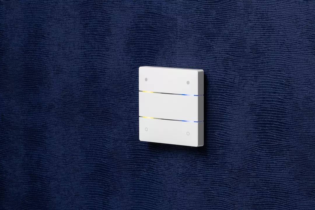 Weißer Lichtschalter mit drei horizontalen Streifen, montiert auf einer dunkelblauen Wand. Schalter zeigt gelbes und blaues Licht an.