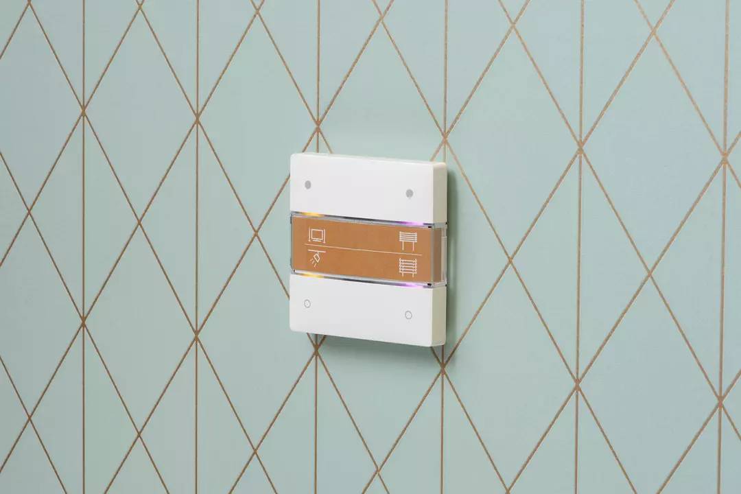 Ein weißer Schalter an einer türkis- und goldfarbenen Wand mit geometrischem Muster. Der Schalter hat drei Symbole: Fernseher, Licht und Heizung.