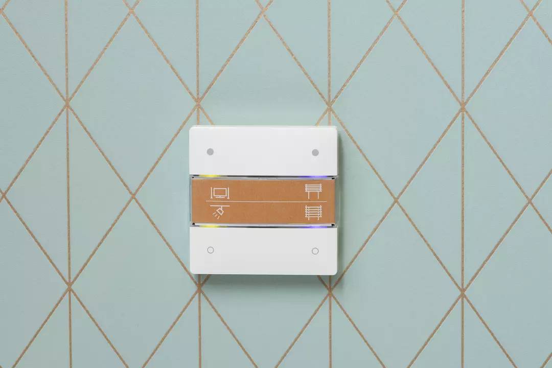 Ein rechteckiger Lichtschalter an einer hellblauen, rautenförmigen Kachelwand mit Symbolen für Beleuchtung und Heizungen.