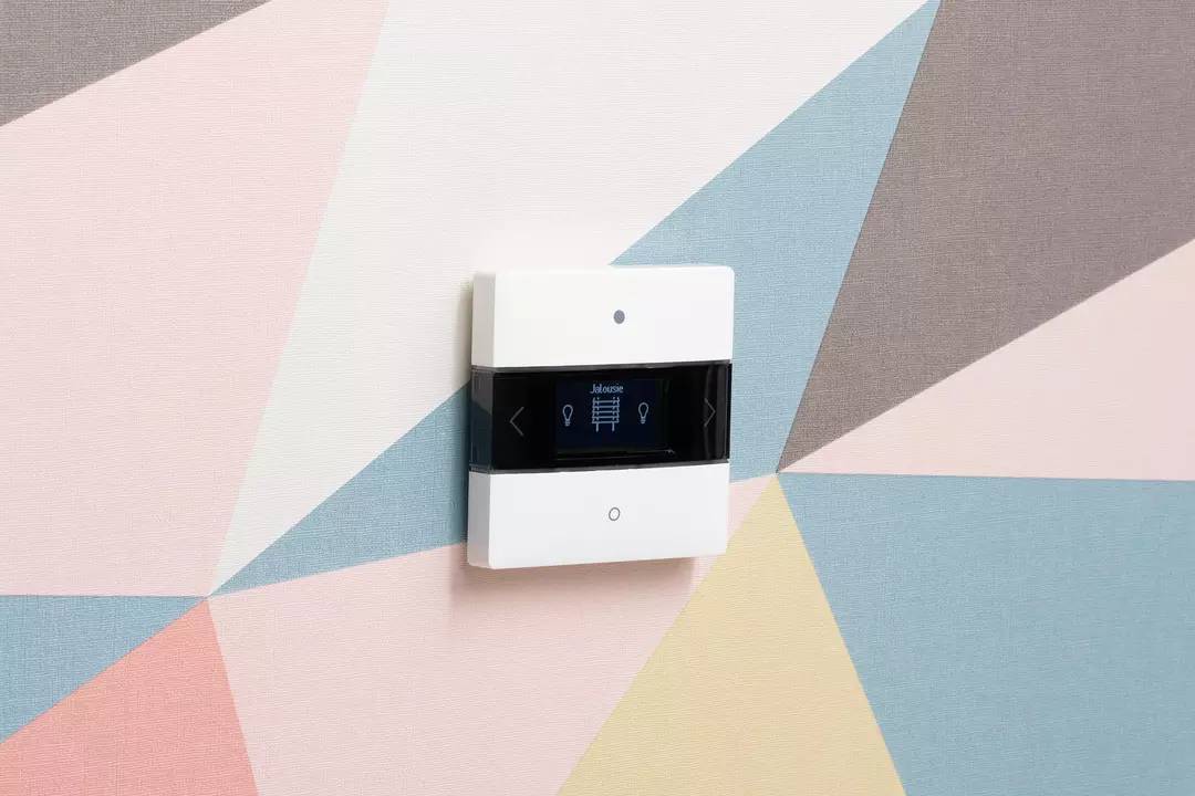 Ein Thermostat an einer wand mit geometrischem Muster. Auf dem Display steht 'Manuell', daneben Pfeiltasten für Einstellungen.