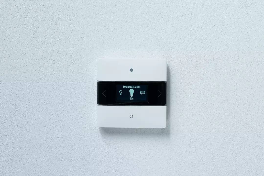Ein weißer Lichtschalter mit digitalem Display an der Wand zeigt 'Deckenleuchte Ein' und Symbole für Lichtsteuerung.