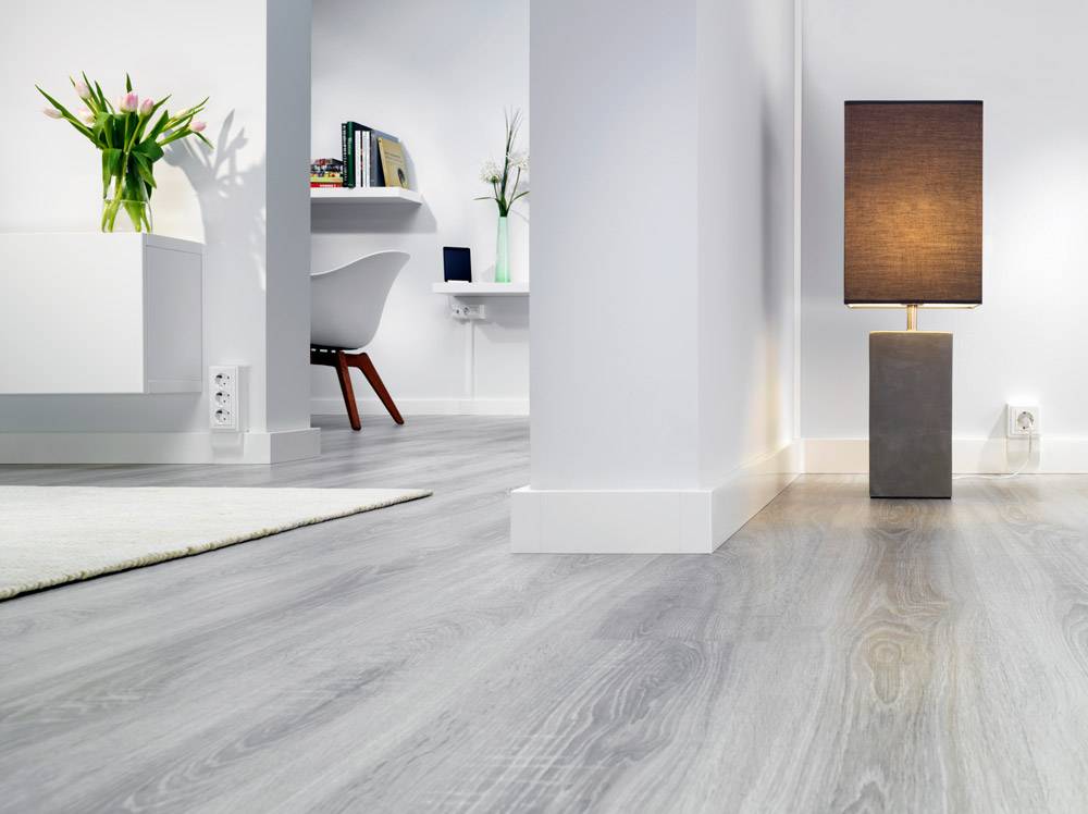 Heller Wohnraum mit grauem Holzboden, Stehlampe und einem modernen Schreibtisch im Hintergrund. Einfache und elegante Einrichtung.