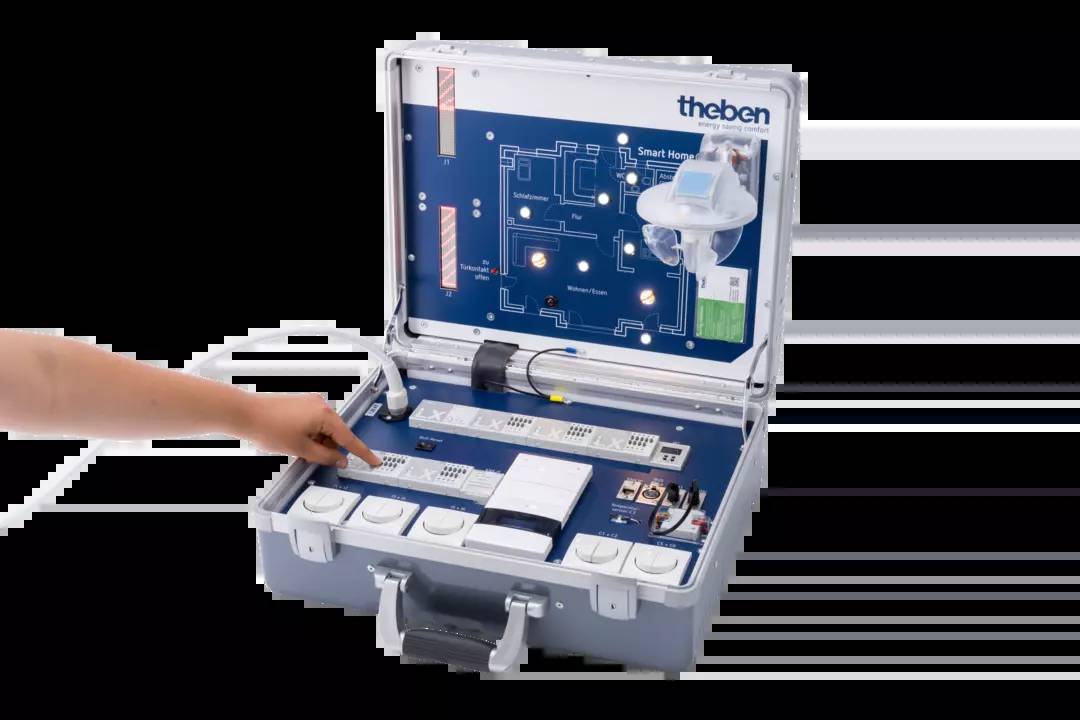 Ein geöffnetes Smart Home-Demokoffer zeigt Schalter, Sensoren und ein kleines Display mit einem Schema eines vernetzten Hauses. Eine Hand zeigt auf die Schalter.