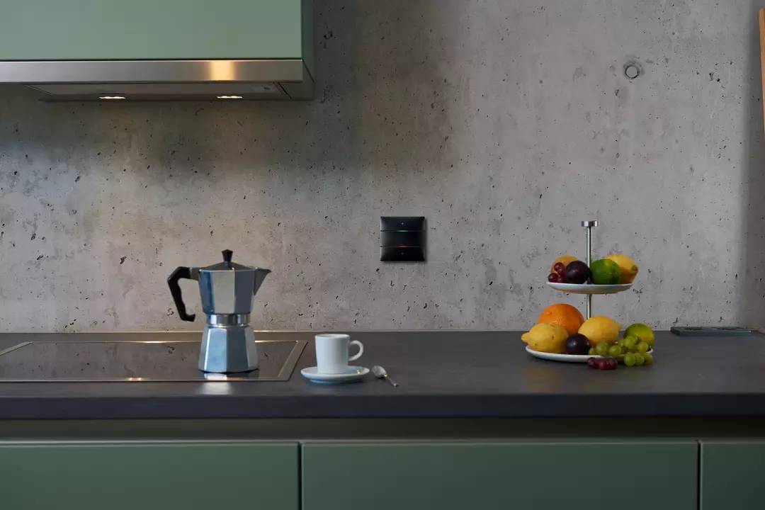 Eine Espressokanne auf einer Küchenarbeitsplatte neben einer Tasse und Obstschale mit Äpfeln, Zitronen und Trauben.