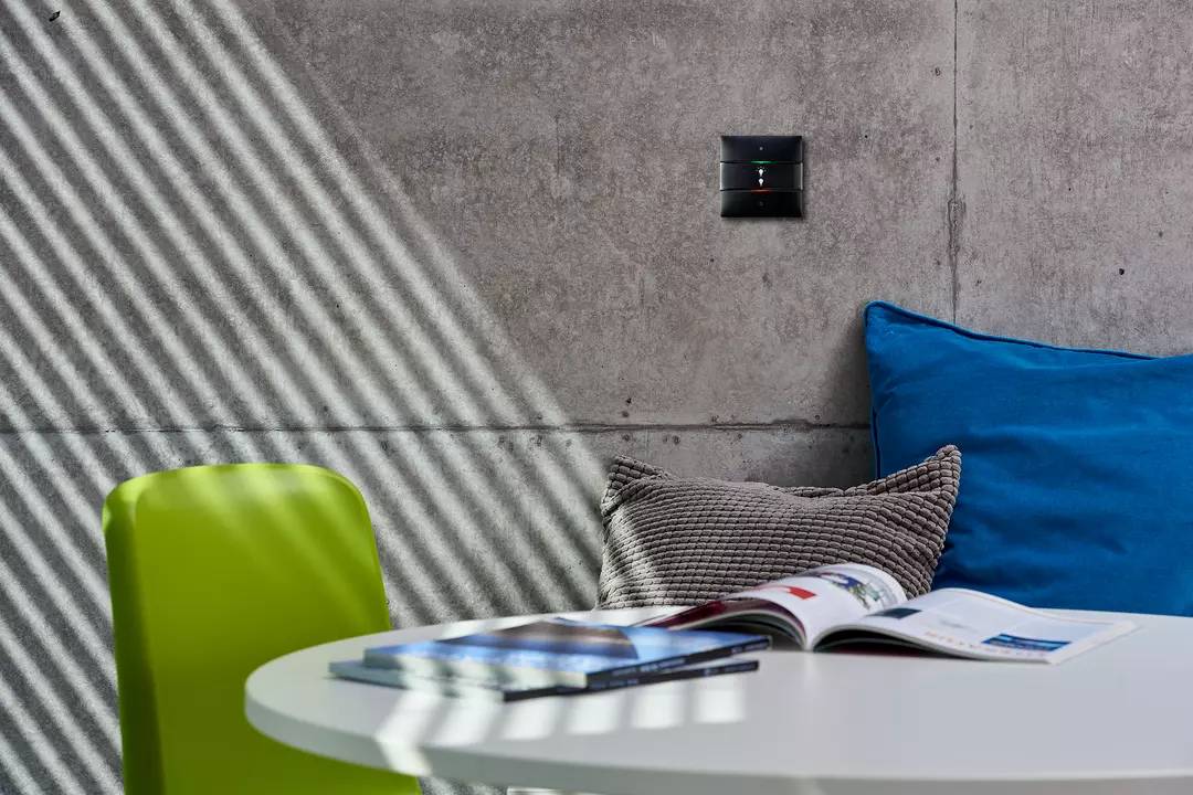 Ein Tisch mit geöffnetem Buch in modernem Raum. Links ein grüner Stuhl, hinten ein blaues Kissen auf einer grauen Betonwand. Schlichtes Ambiente.