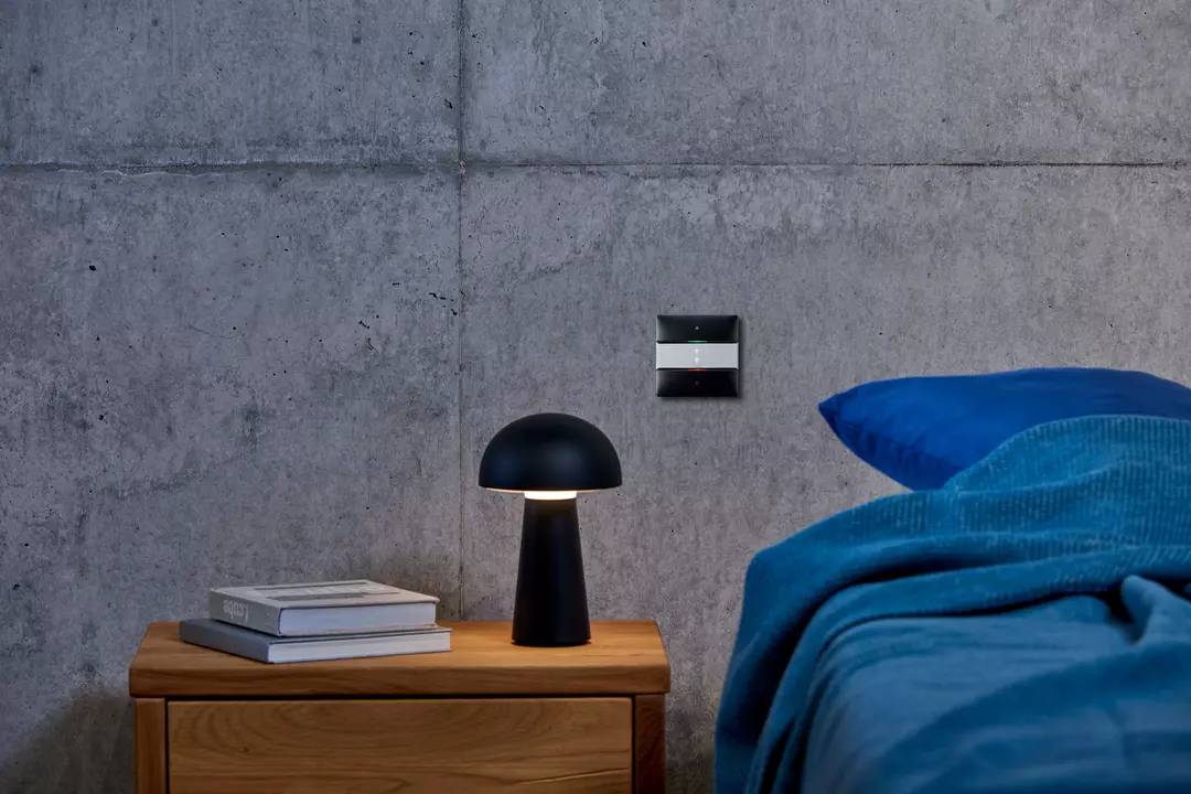 Ein modernes Schlafzimmer zeigt eine blau bezogene Decke auf einem Bett, einen Holztisch mit schwarzer Lampe und eine Betonwand.