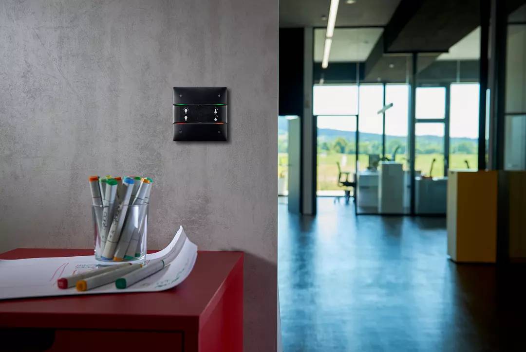 Ein moderner Büroraum mit großen Fenstern, rotem Tisch und einem Glas mit farbigen Stiften im Vordergrund; Lichtschalter an der Wand.