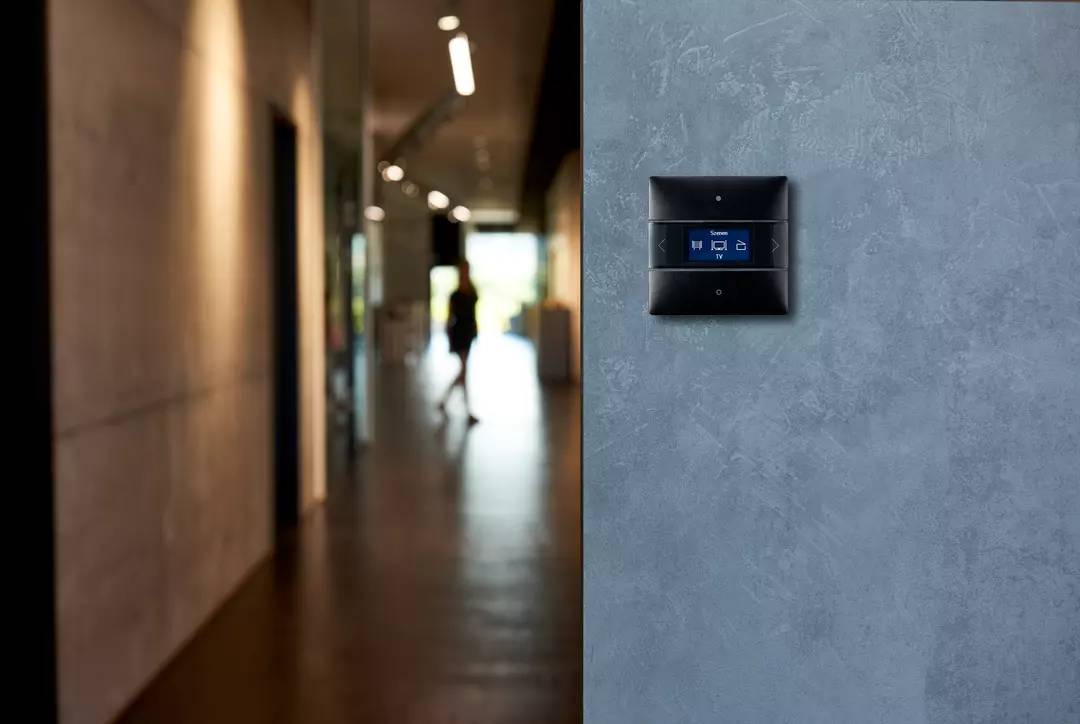 Ein moderner Lichtschalter mit digitalem Display an einer Betonwand, im Hintergrund ein unscharfer Flur mit einer Person.
