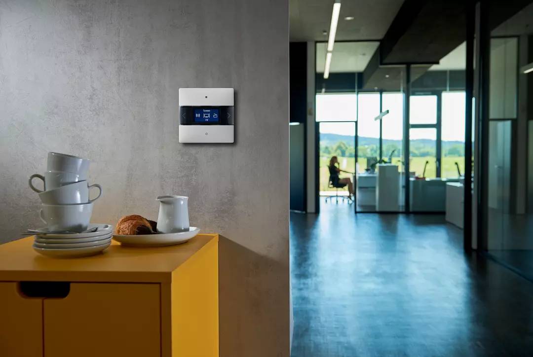 Ein modernes Büro mit einem digitalen Thermostat an der Wand. Im Hintergrund sitzt eine Person an einem Tisch mit Blick auf eine Landschaft.