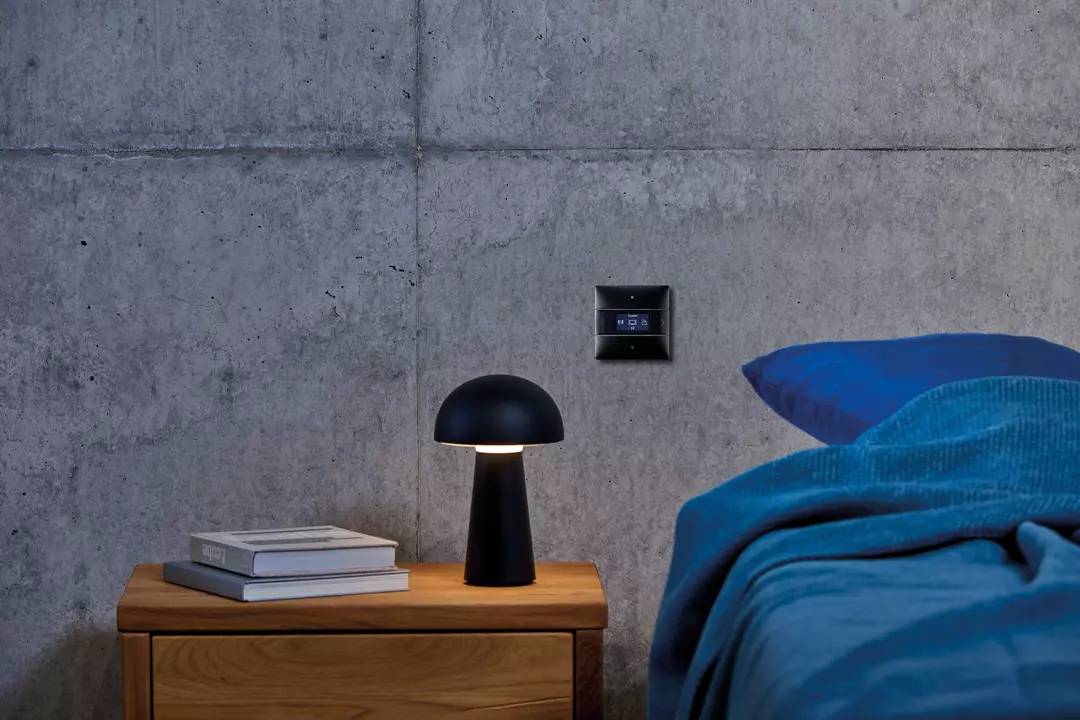 Nachtischlampe auf Holztisch neben einem Bett mit blauer Decke, an Betonwand. Dunkles, gemütliches Schlafzimmerambiente.