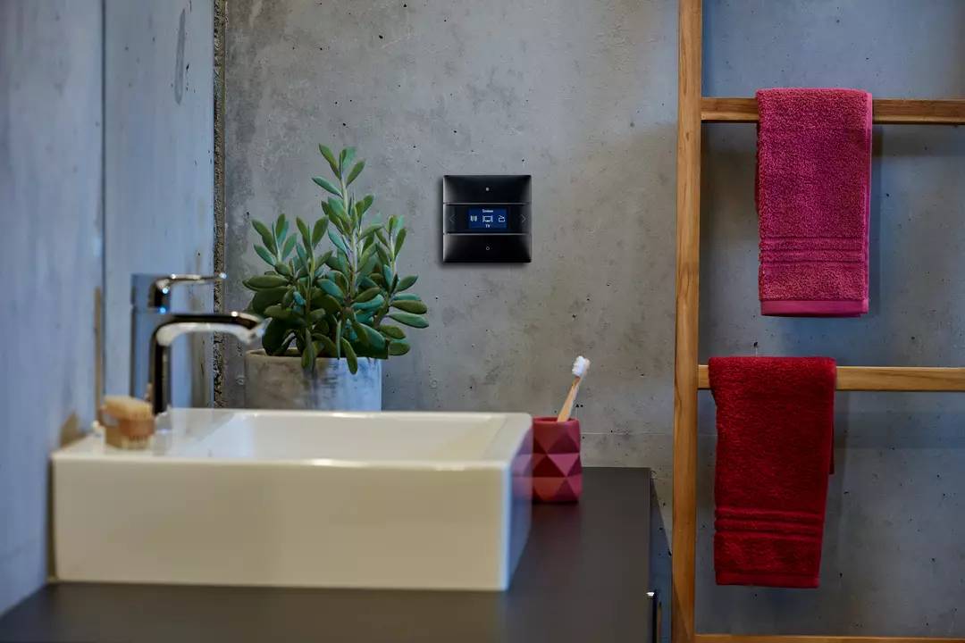 Modernes Badezimmer mit grauer Wand, Pflanze neben Waschbecken, digitalem Steuerungsgerät an der Wand; rote Handtücher auf Holzhalter.