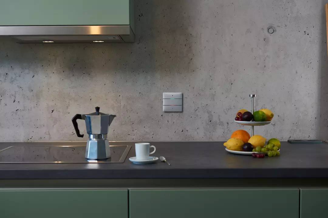 Küche mit Espresso-Maschine auf Herd, Tasse auf Arbeitsplatte und Obstteller rechts. Schlichte, moderne Einrichtung.