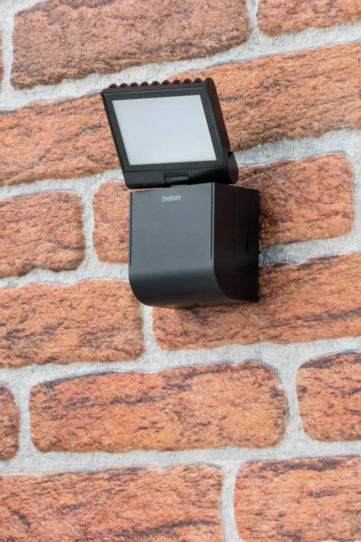 Ein schwarzer Bewegungssensor mit Solarzelle ist an einer ziegelroten Wand montiert, nach vorne geneigt zur Erfassung von Bewegungen.