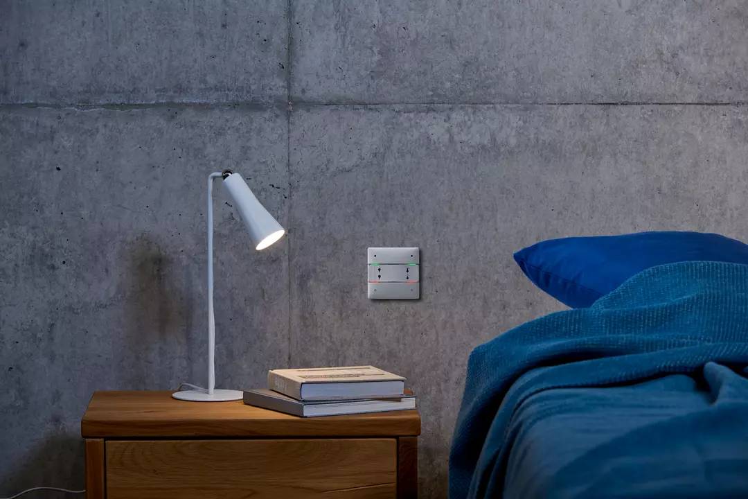 Nachttisch mit weißer Lampe, zwei Büchern und einer Betonwand im Hintergrund. Rechte Seite eines blauen Bettes ist sichtbar.