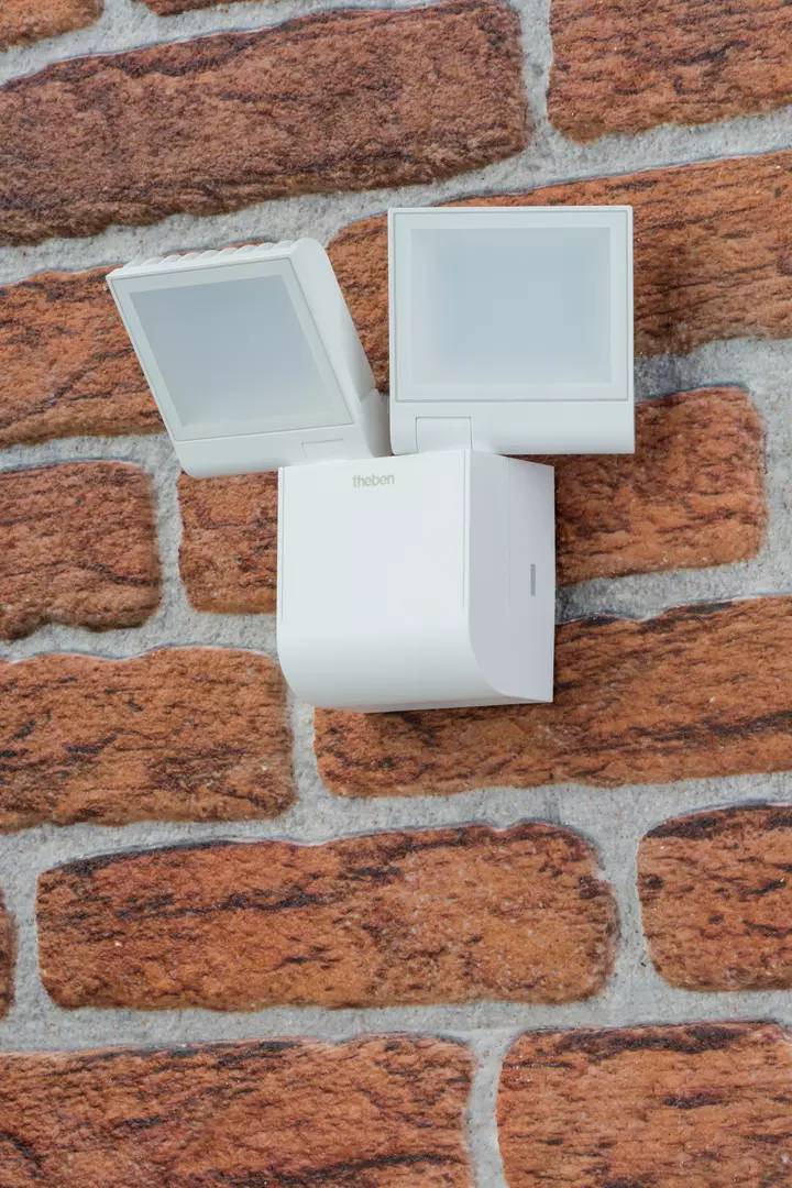 Ein weißer Bewegungsmelder ist an einer Ziegelwand montiert. Der Detektor hat zwei rechteckige Sensoren für die Bewegungserkennung.