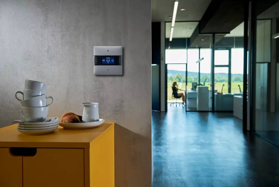 Ein moderner, heller Büroraum mit großen Fenstern zeigt eine Person im Hintergrund. Im Vordergrund ist ein gelber Tisch mit Frühstücksgeschirr.