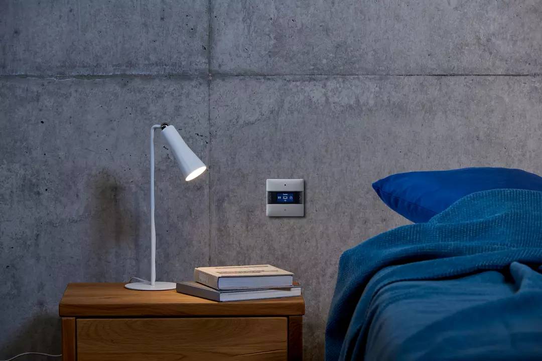 Ein modernes Schlafzimmer mit einer Tischlampe auf einem Holztisch, einem intelligenten Thermostat an der Wand und einem Bett mit blauer Bettwäsche.