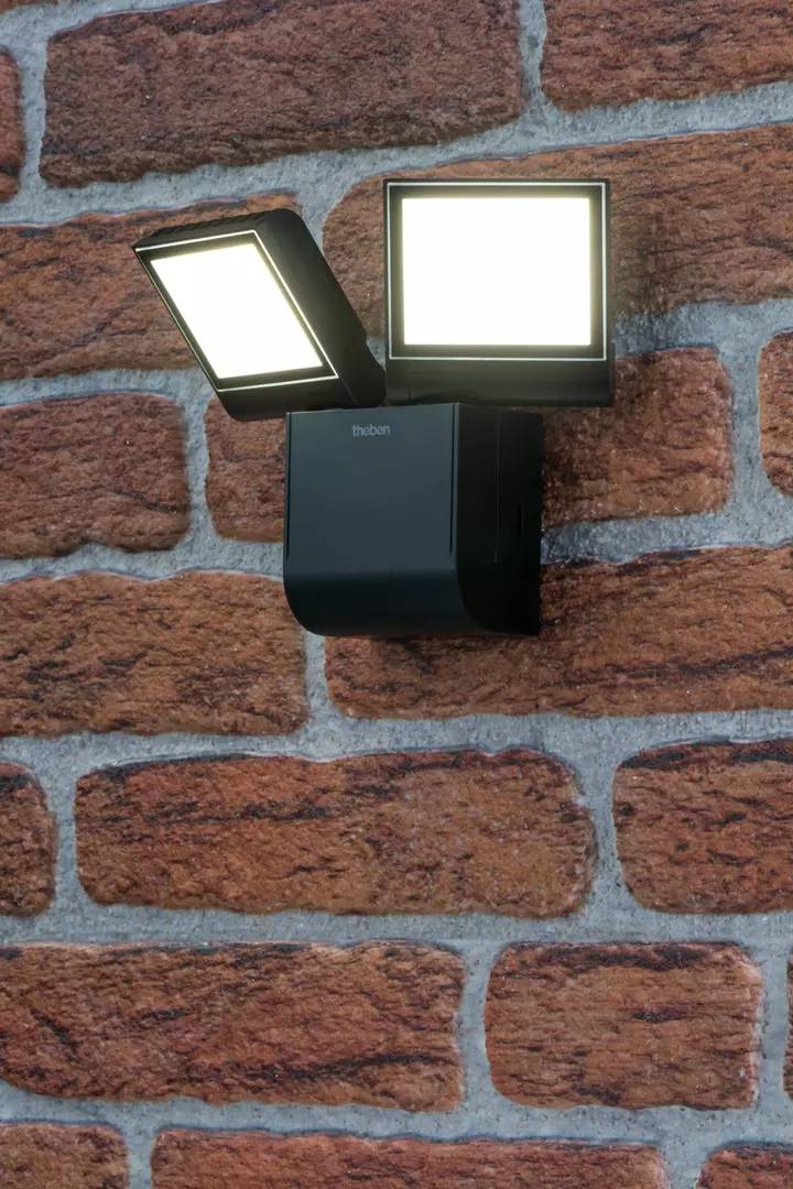 Eine zweilampige, schwarze Wandleuchte leuchtet auf eine Backsteinmauer. Sie wirft helles Licht und hebt die Textur der Ziegel hervor.