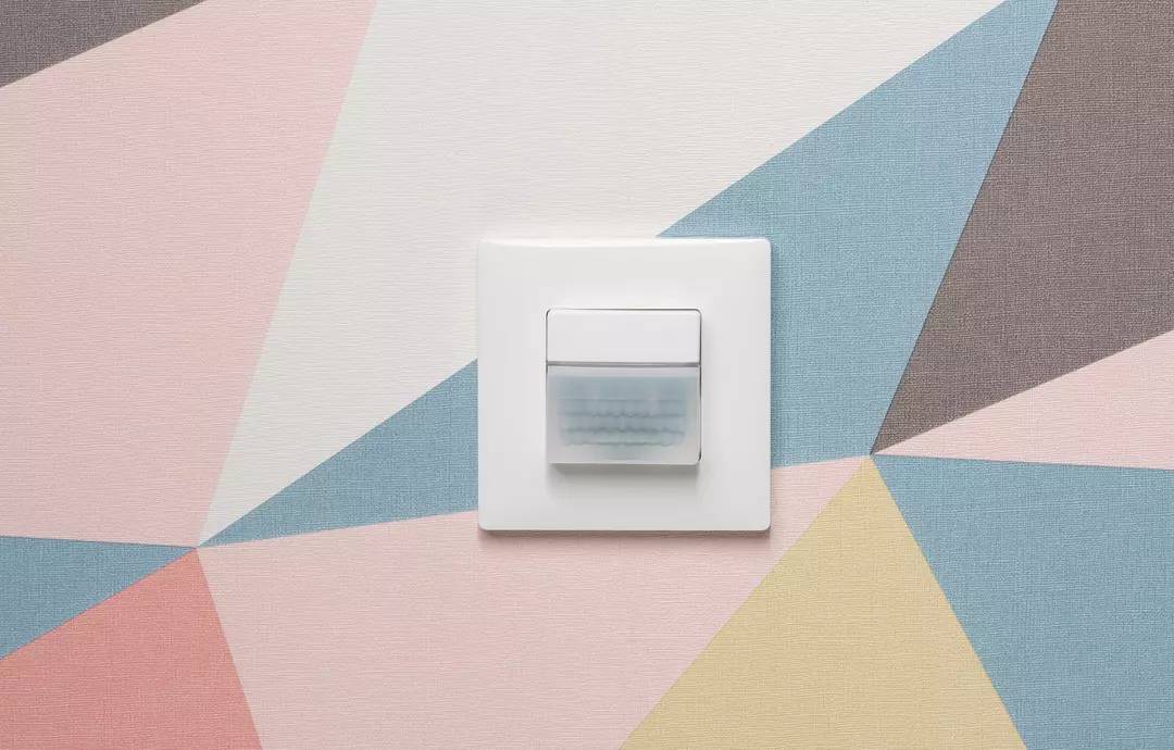 Ein weißer Lichtschalter mit Bewegungsmelder an einer bunt gemusterten Wand. Der Schalter ist zentral platziert.