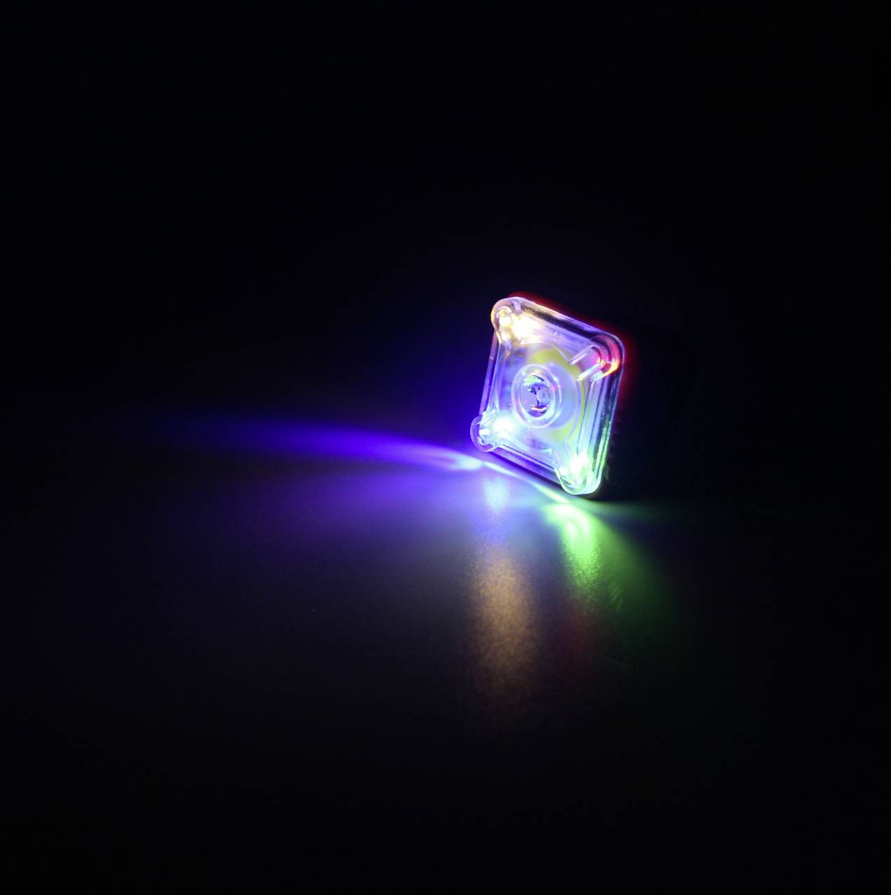 Ein kleines, quadratisches LED-Licht in verschiedenen Farben leuchtet in der Dunkelheit. Es erzeugt einen Regenbogen-Effekt auf der Oberfläche.