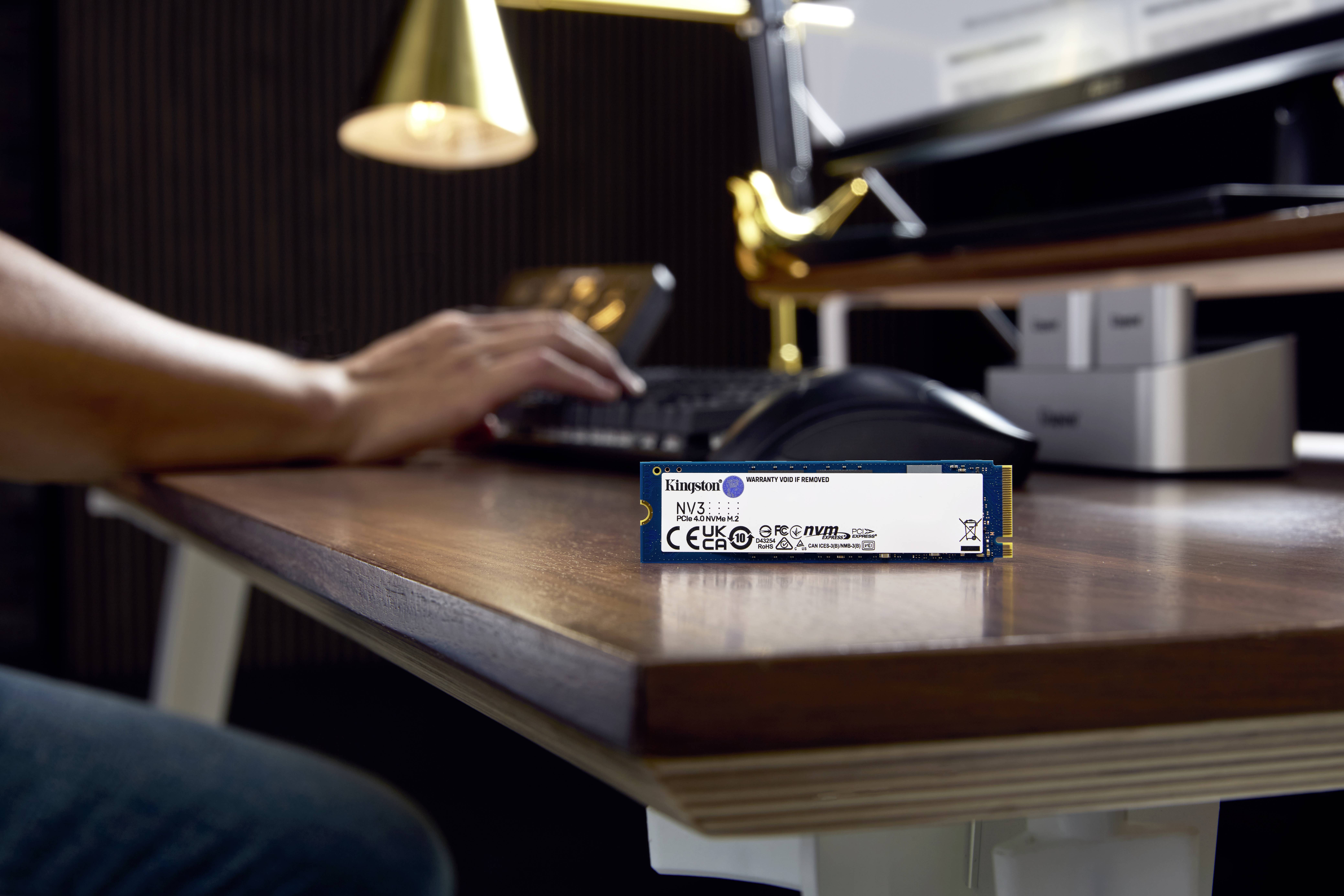 Eine Person arbeitet am Computer. Im Vordergrund auf dem Tisch liegt ein SSD-Modul von Kingston mit einem markanten blauen Etikett.