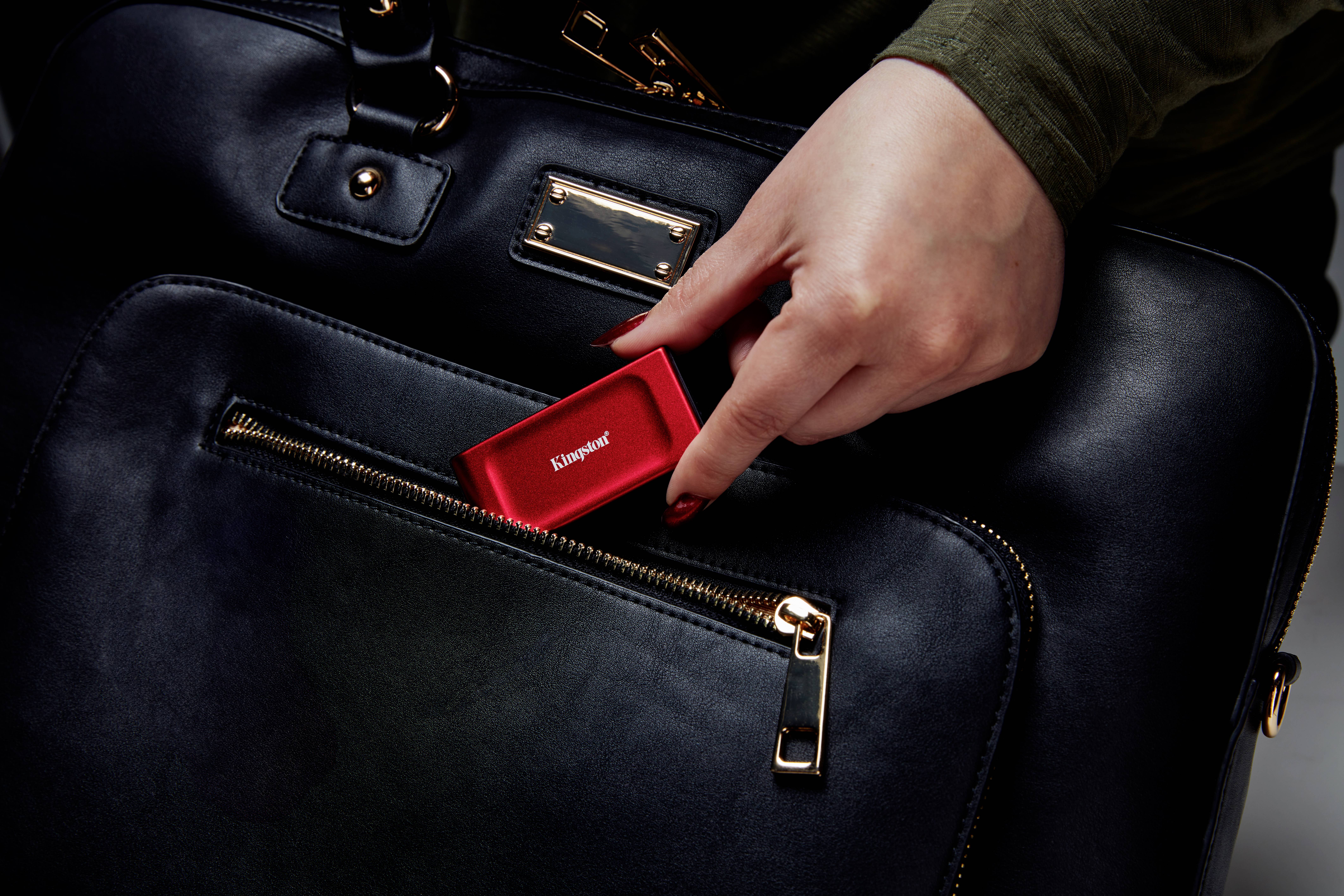 Eine Hand steckt ein rotes tragbares Gerät in die vordere Tasche einer schwarzen Ledertasche.