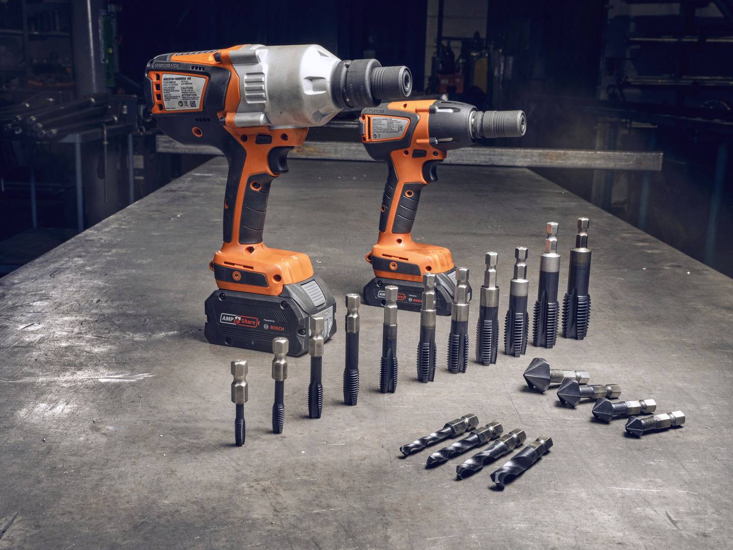 Zwei orange Akkuschrauber mit Zubehör auf einem Werkstatttisch. Links ist der größere Schrauber, rechts der kleinere. Verschiedene Bohr- und Schraubaufsätze vorne angeordnet.