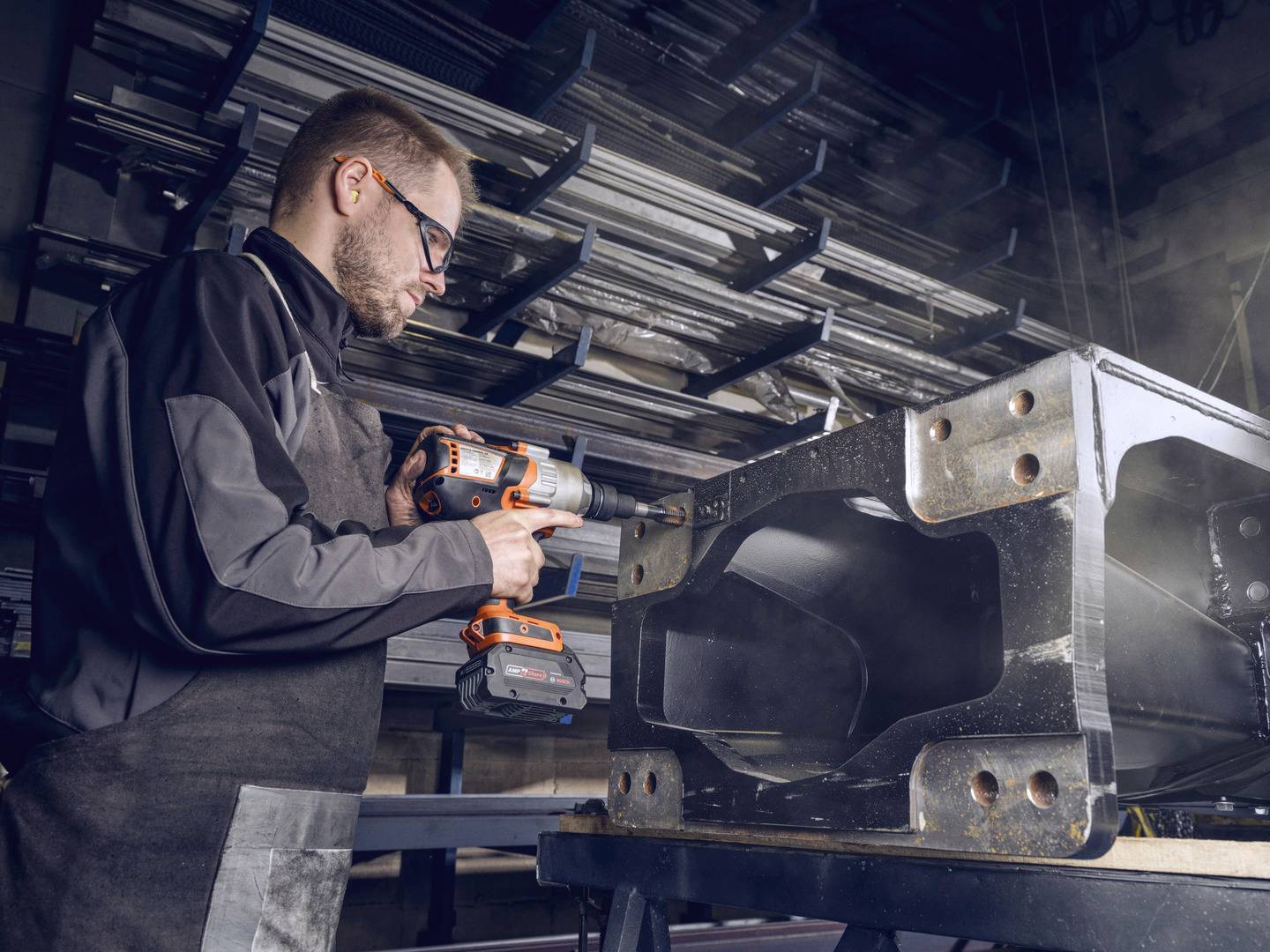Ein Mann in Arbeitskleidung benutzt eine Bohrmaschine, um an einem großen Metallrahmen zu arbeiten. Im Hintergrund sind Regale mit Metallteilen.