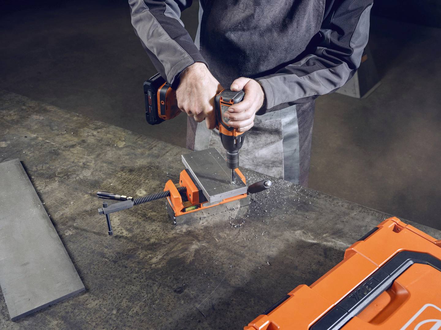 Eine Person in Arbeitskleidung verwendet eine orange Bohrmaschine, um ein Metallstück in einer Halterung auf einem Tisch zu bearbeiten.