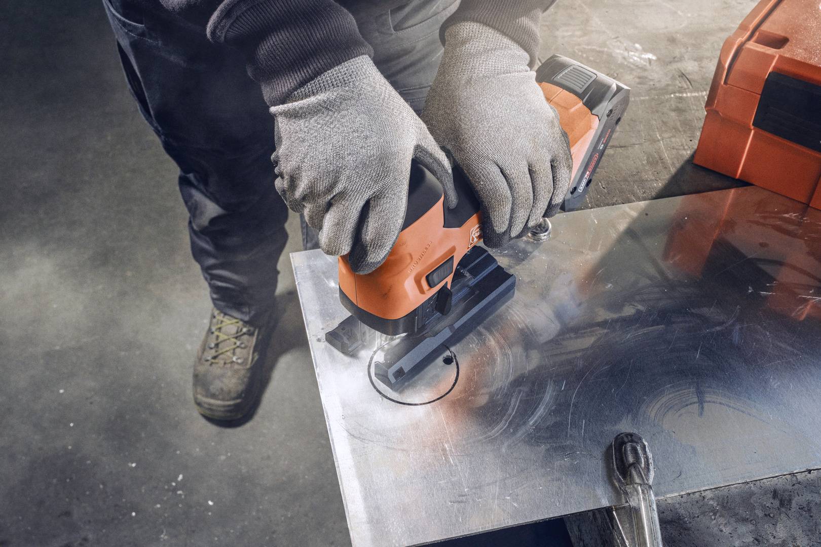 Eine Person schneidet mit einer Stichsäge ein rundes Loch in ein Metallblech. Sie trägt Arbeitshandschuhe und Stiefel.