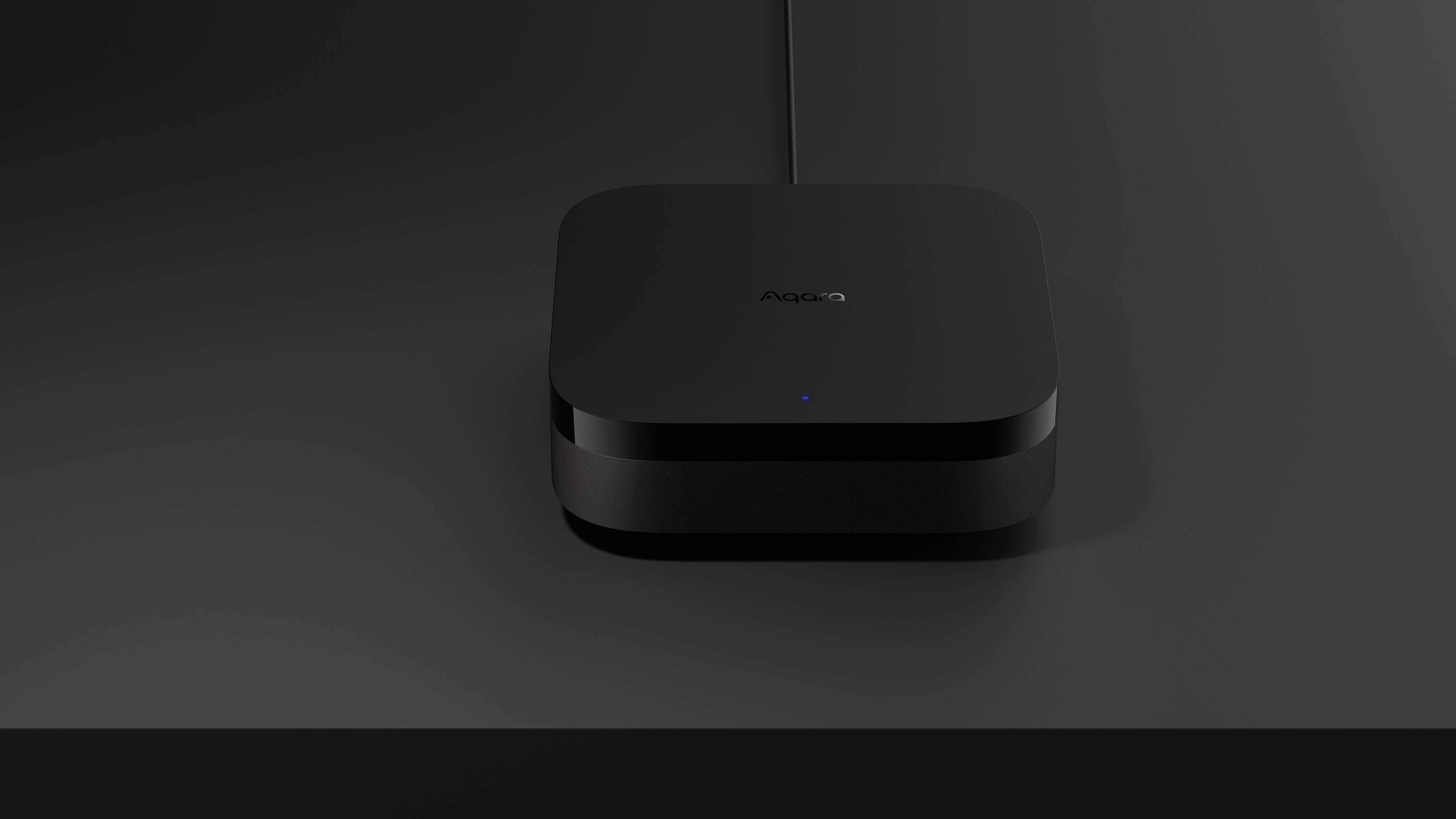 Ein schwarzes, quadratisches Gerät mit abgerundeten Ecken und einem leuchtenden blauen Punkt steht auf einem dunklen Tisch.