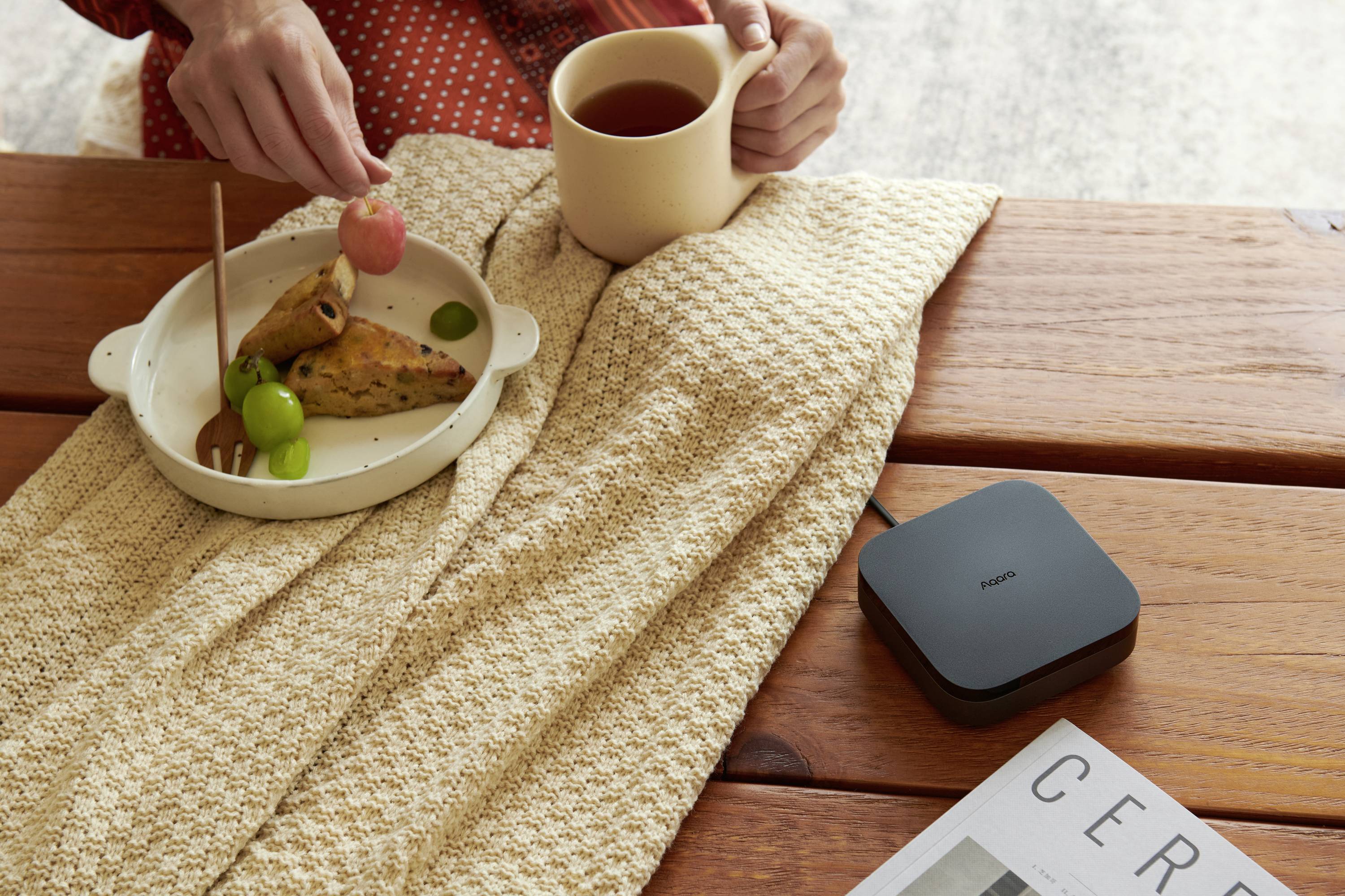 Eine Person genießt Tee mit einer kleinen Mahlzeit aus Gebäck und Trauben. Daneben liegt ein elektronisches Gerät und eine Zeitschrift.