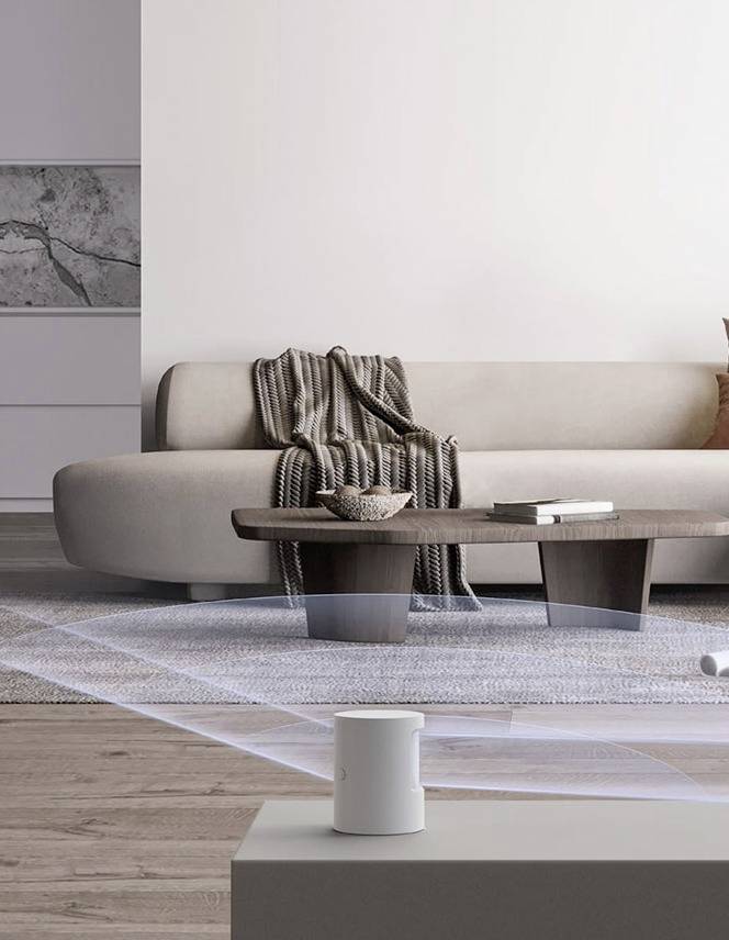 Ein modernes Wohnzimmer mit einem cremefarbenen Sofa, einem Holztisch und einer gestreiften Decke. Im Vordergrund steht ein weißer Lautsprecher.