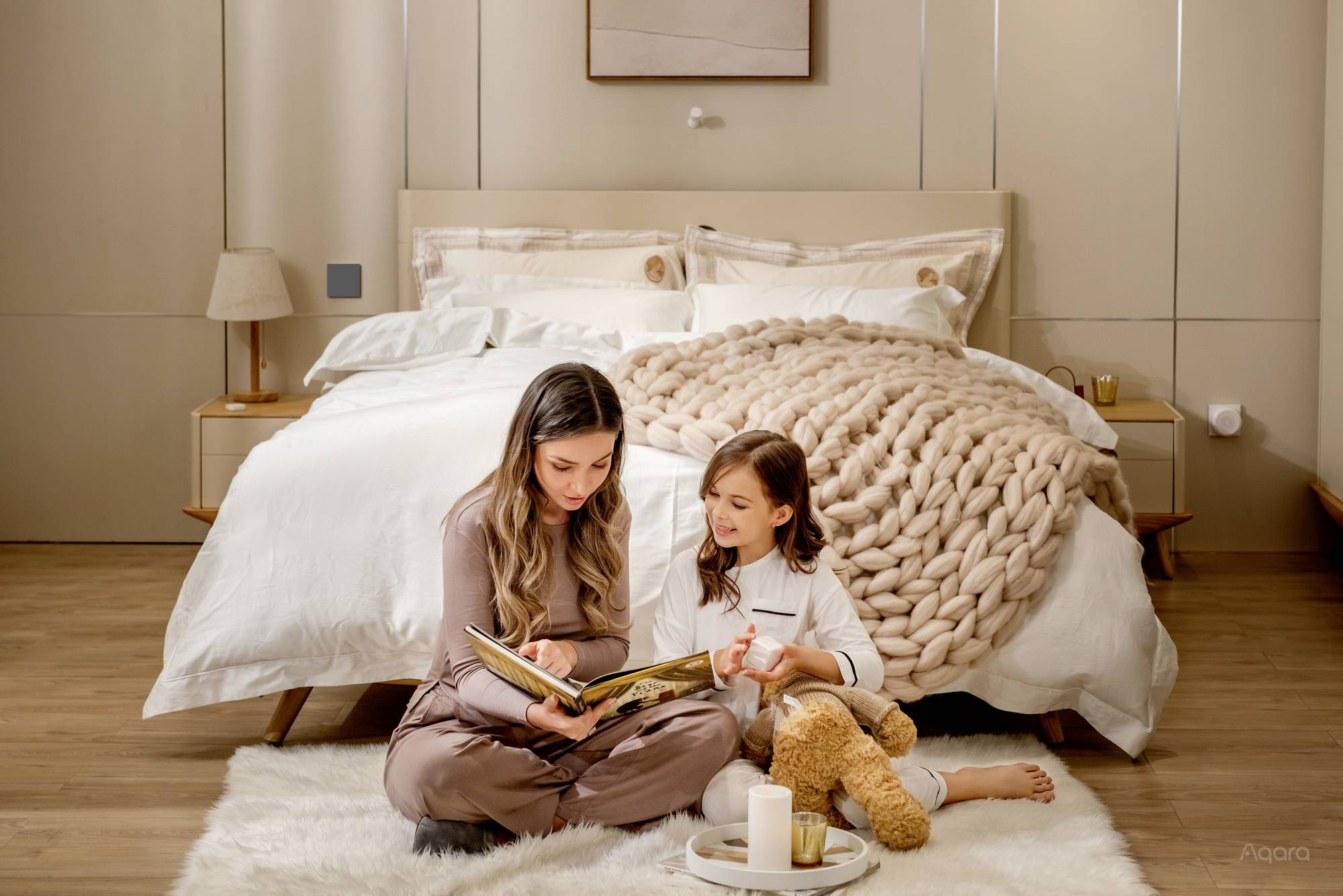 Eine Frau und ein Mädchen sitzen auf einem Teppich vor einem Bett und lesen ein Buch zusammen. Das Mädchen hält einen Plüschbären.