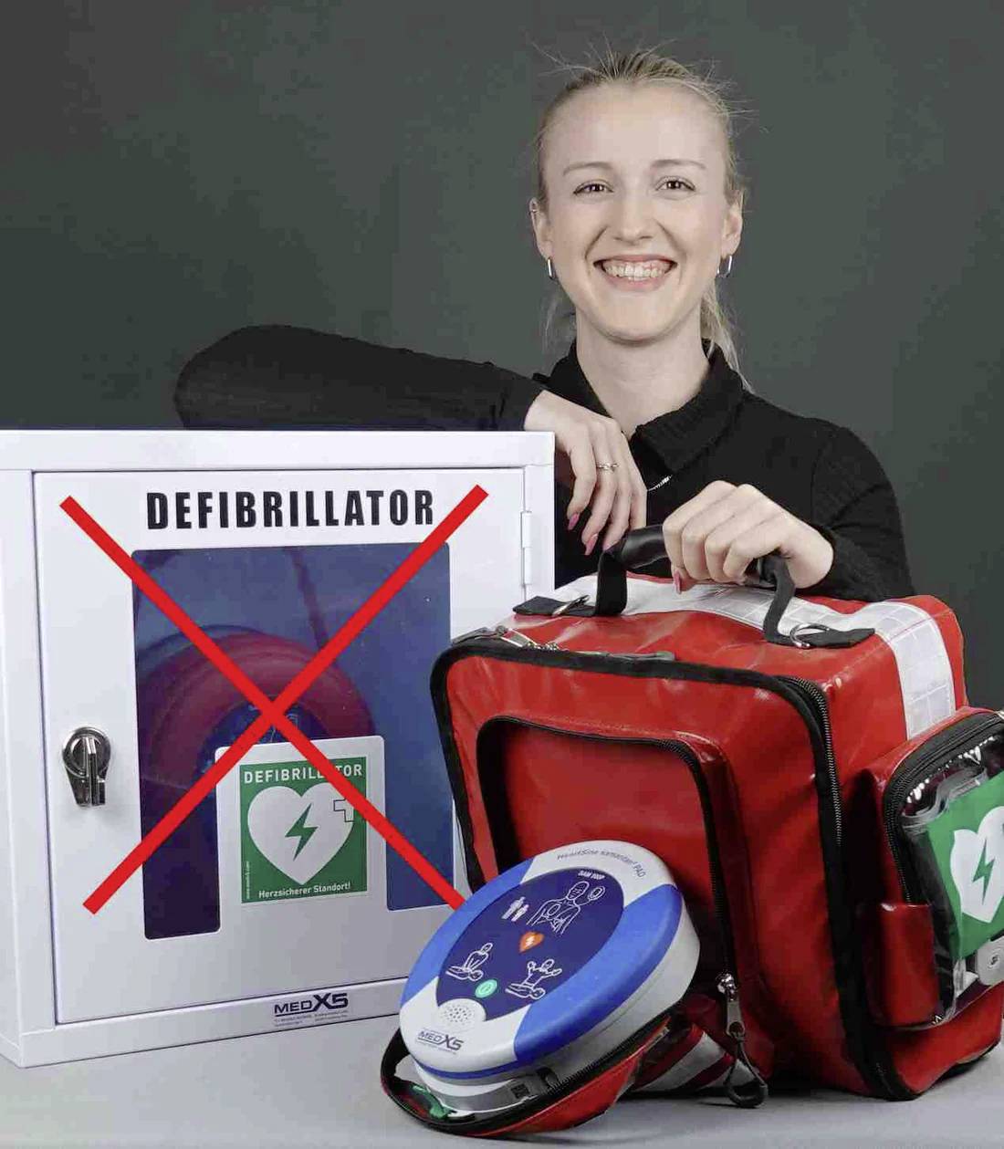 Eine Person lächelt neben einem Defibrillator-Schrank, der mit einem roten X markiert ist, und hält eine rote Notfalltasche mit einem tragbaren Defibrillator.