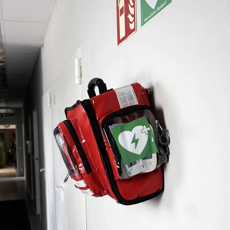 Ein Erste-Hilfe-Rucksack mit AED-Symbol hängt an der Wand unter einem Feuerlöscher-Schild in einem hellen Flur.